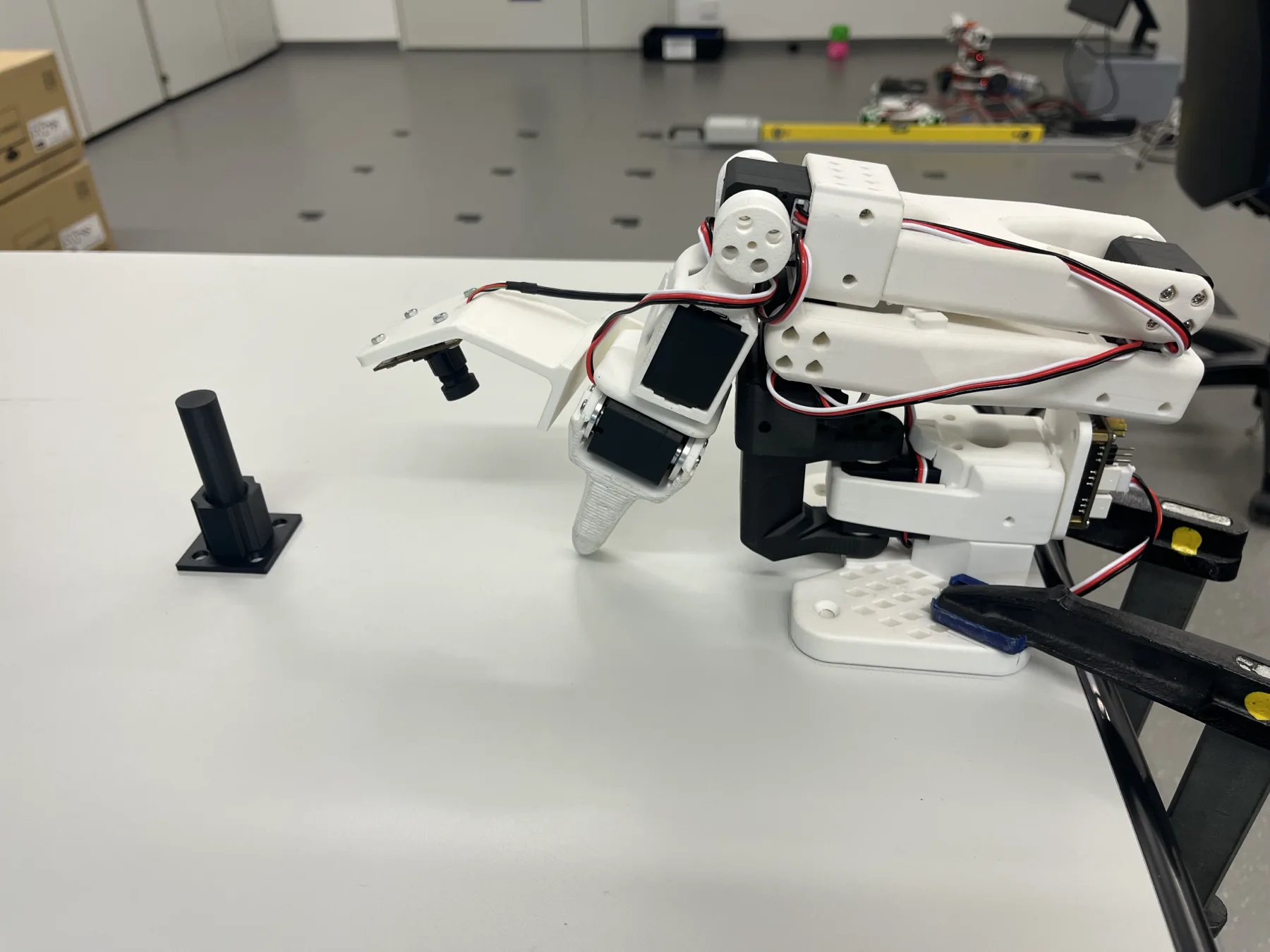 The SO-101 arm with its wrist camera on a white table, with the black peg fixture in front of it